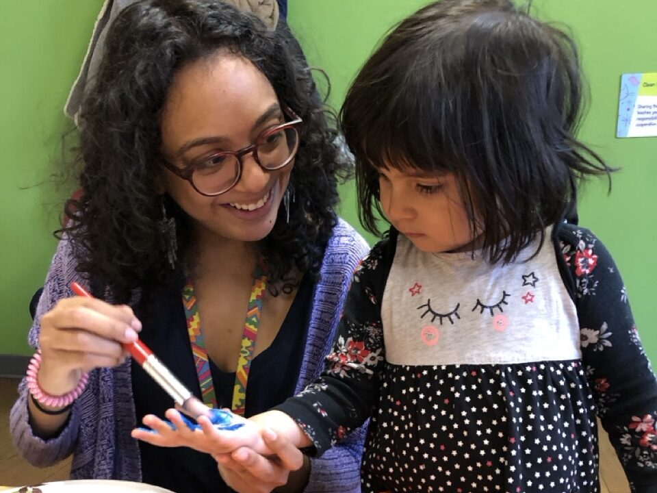 An adult paints on a child's hand, while the child watches.