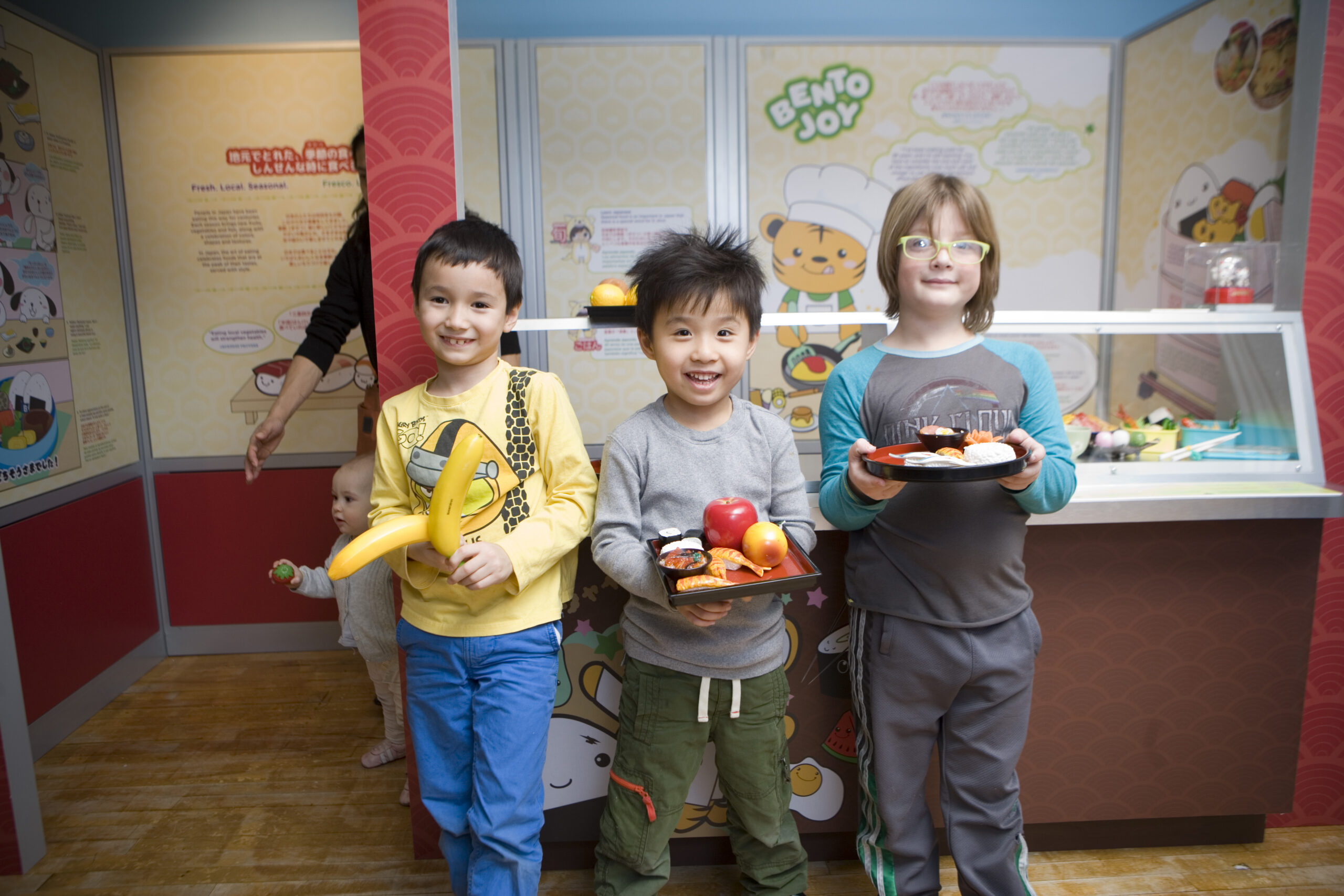 Three boys smile in font of a sushi bar with trays of Japanese food in the Hello from Japan exhibit.