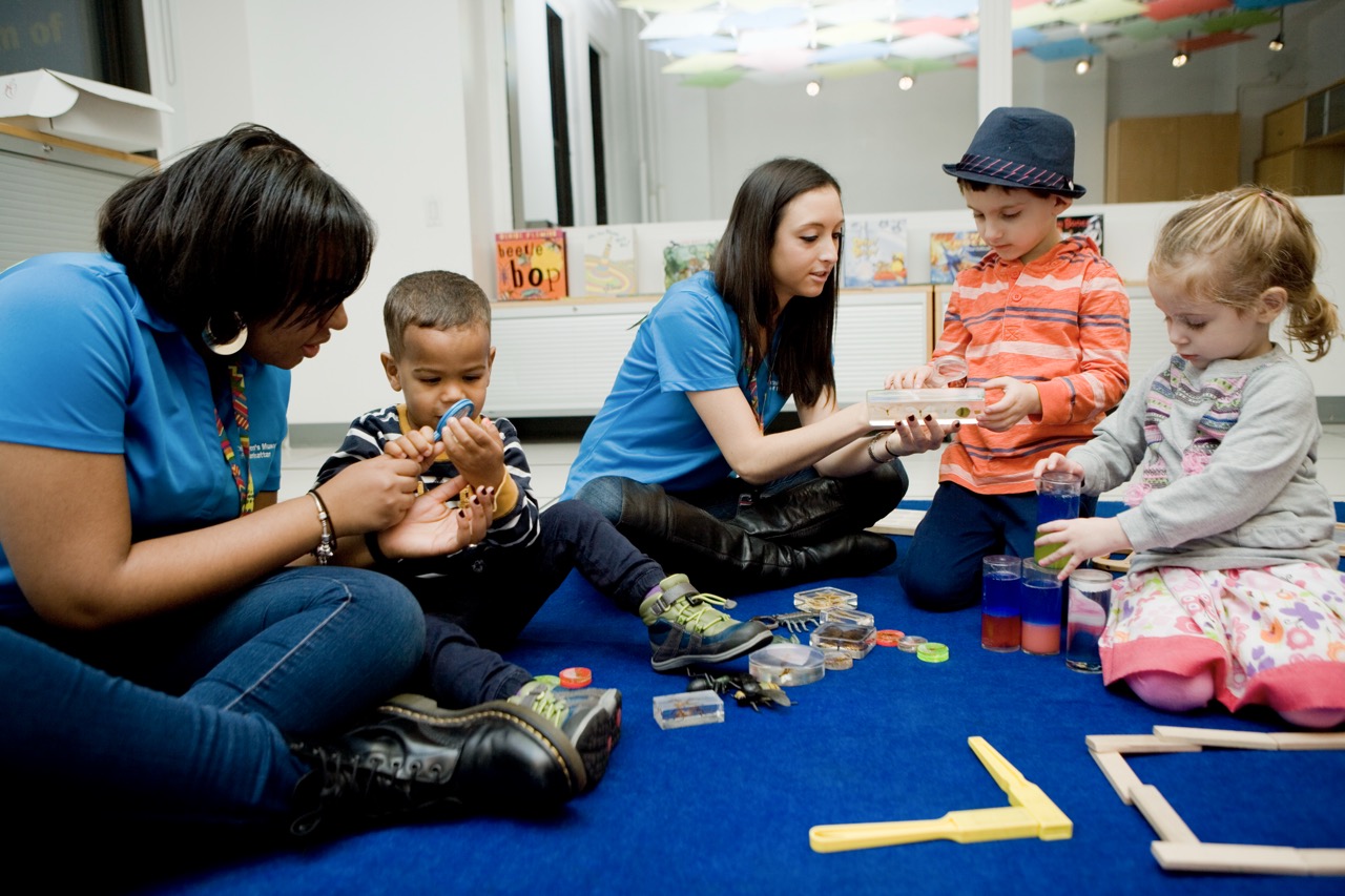 Two adults sit on the floor with three small children, who are investigating small objects.