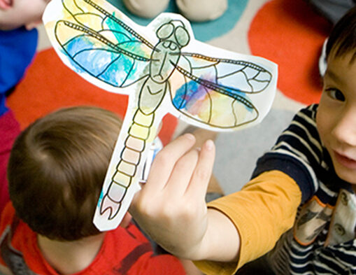 A child holds up a painted dragonfly illustration.