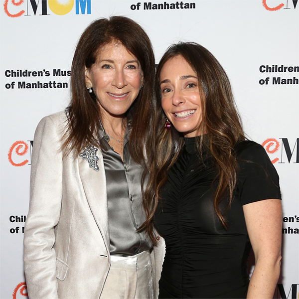 Two woman smile at the camera in front of a step-and-repeat with CMOM's logo on it
