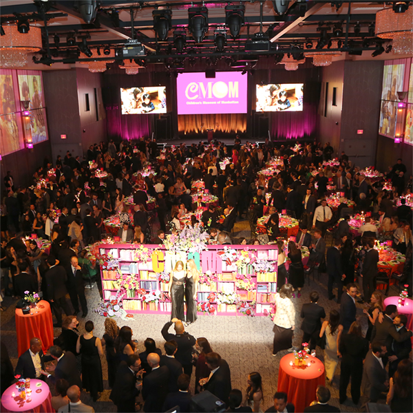 A room with many tables and people for a fancy gala event. CMOM's logo is visible on a screen on the stage at the back of the room.