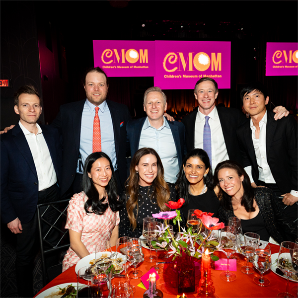 Five adult men stand behind four seated adult women at a gala event. CMOM's logo is seen on screens behind the standing men.