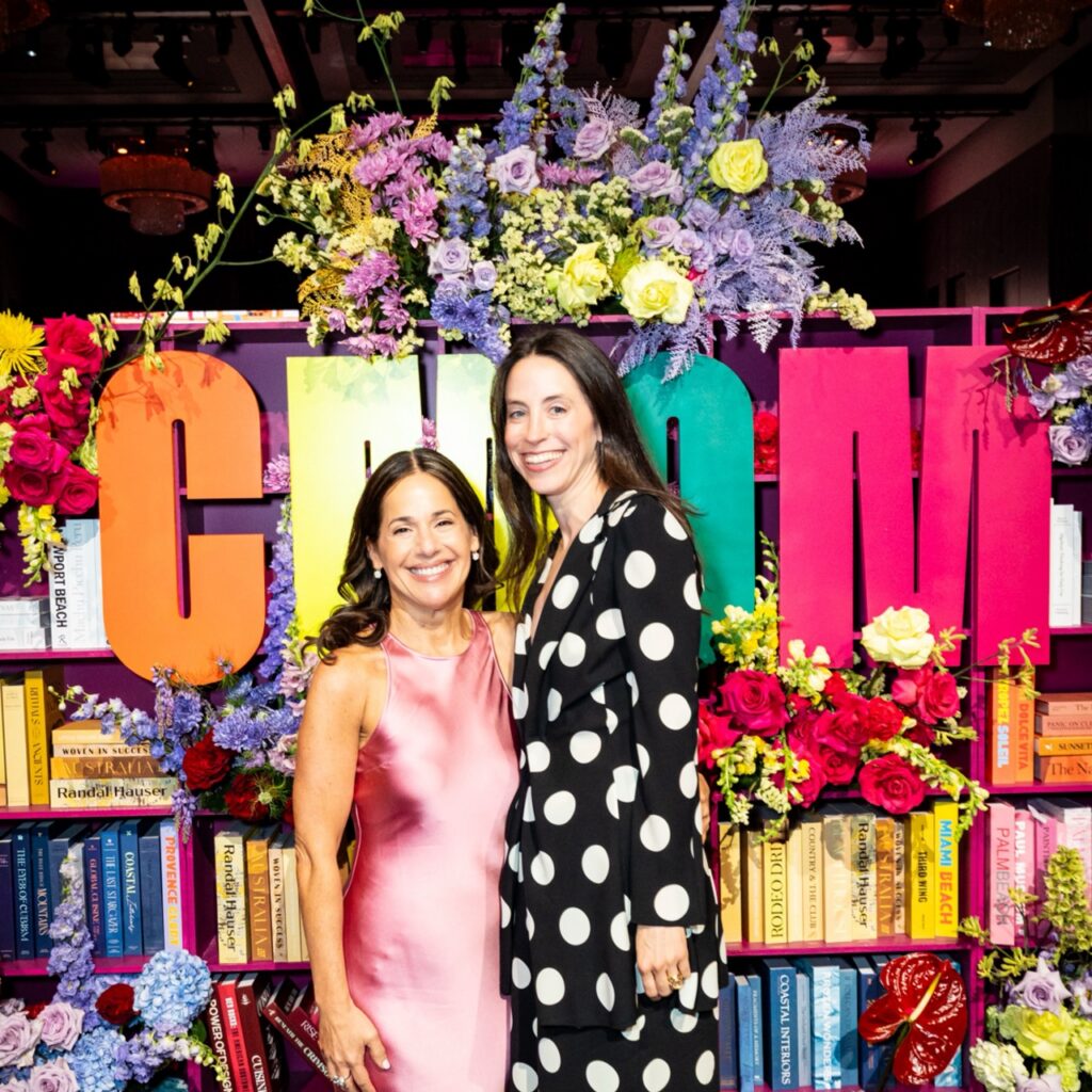 Two adult women smile at the camera in front of a book-and-floral photo opportunity backdrop at a gala.