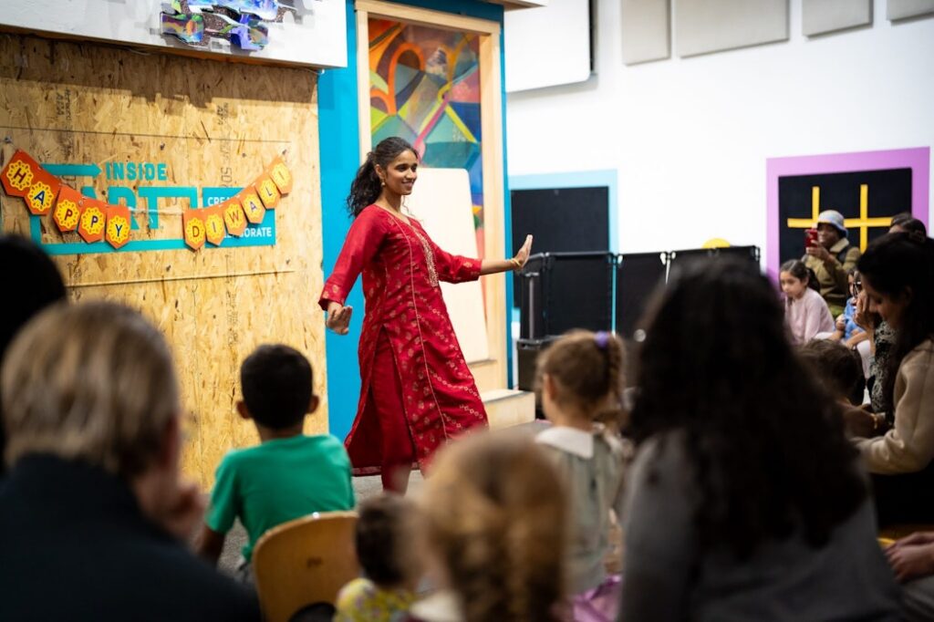 A woman in Indian dress dances in front of a crowd, there is a banner behind her that says "Happy Diwali"