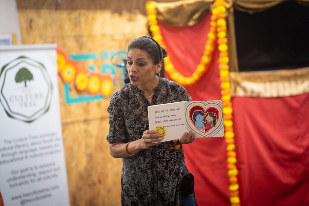 A woman holds a children's book, and appears to be reading it to a crowd. She is wearing a headset microphone.