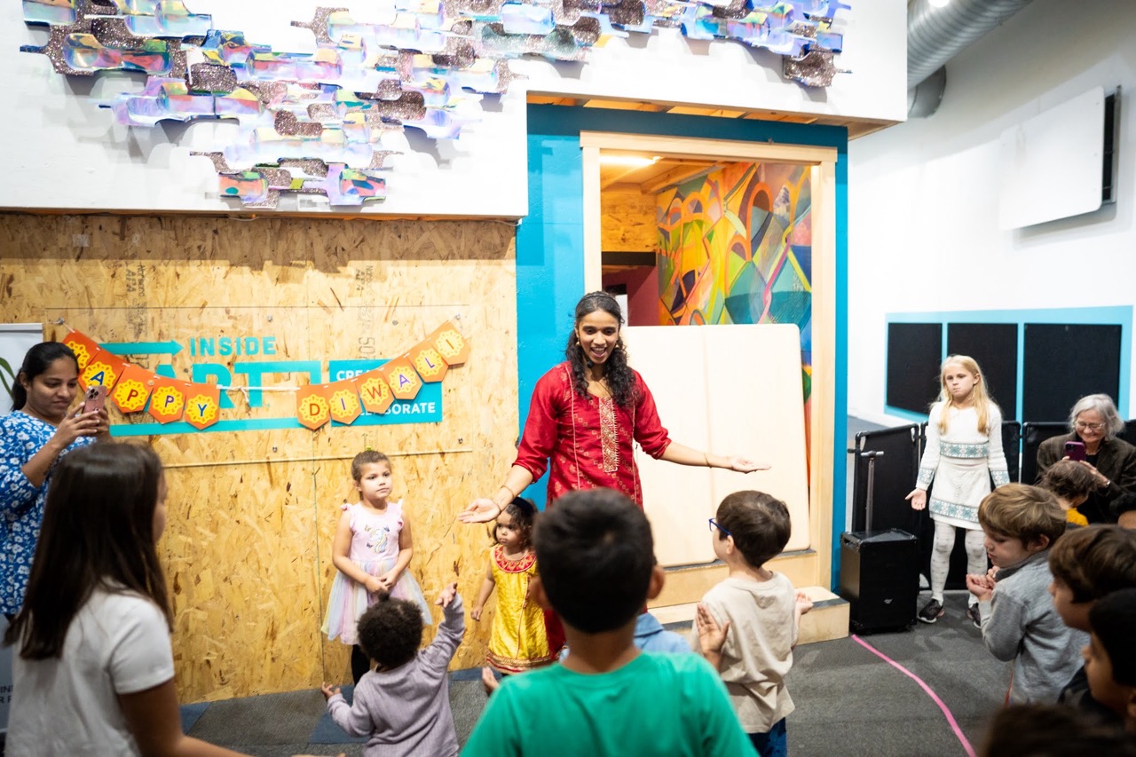 A woman presents to a crowd of children in front of a sign that says "Happy Diwali"