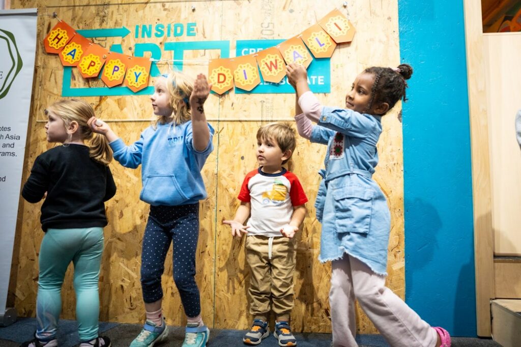 Children look to the left in front of a wooden wall with a banner hanging on it that says "Happy Diwali"