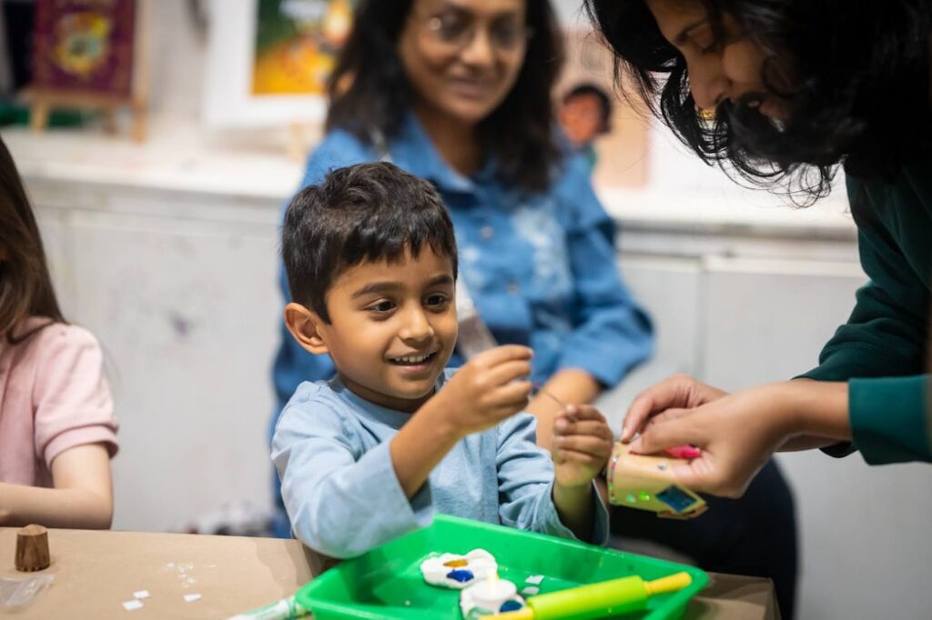 Child smiles while looking at and holding craft supplies. An adult helps him and another adult in the background smiles and watches.