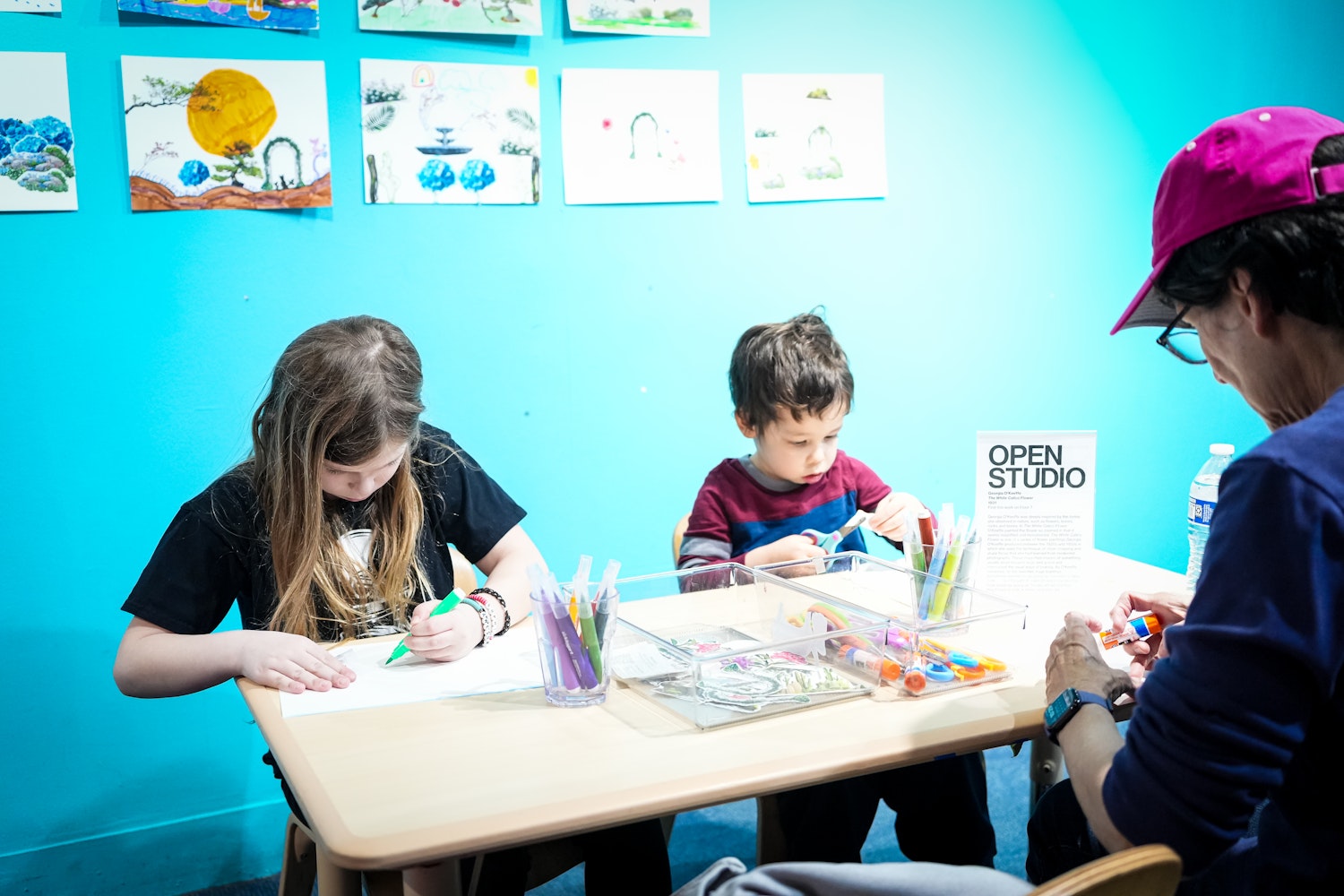 A girl and a boy sit at a table drawing with markers while an adult supervises beside them near an Open Studio sign at the Children’s Museum of Manhattan.